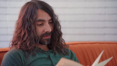 hombre hispano con cabello largo y castaño rizado y barba leyendo felizmente el libro rojo dando vuelta a la página en el sofá naranja