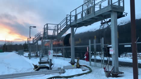 Vista-Panorámica-Izquierda-De-Una-Estación-De-Carga-De-Combustible-En-Un-Depósito-De-Trenes-En-Invierno,-Con-Nieve-En-El-Suelo-Y-Un-Amanecer-En-El-Fondo