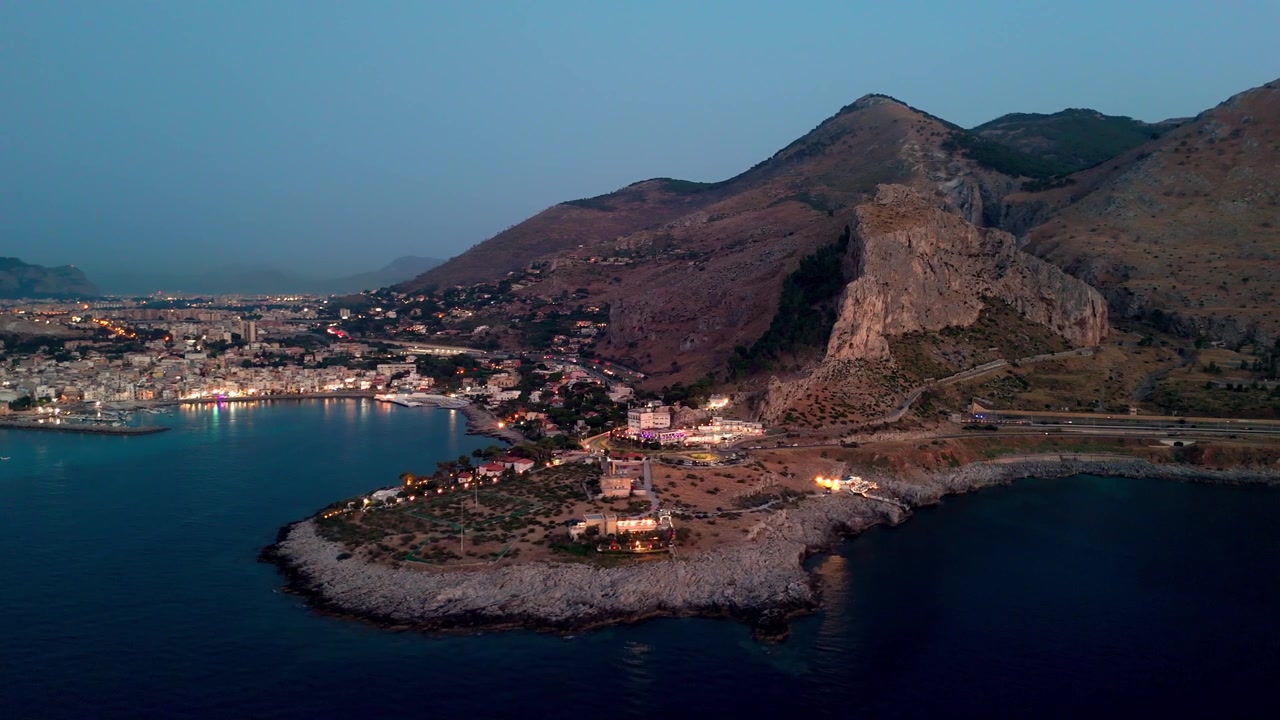 Sferracavallo, Palermo, Sicilya, İtalya'da sahil köyünün, liman ışıklarının ve kayalık kıyı şeridinin havadan mavi saat görünümü.