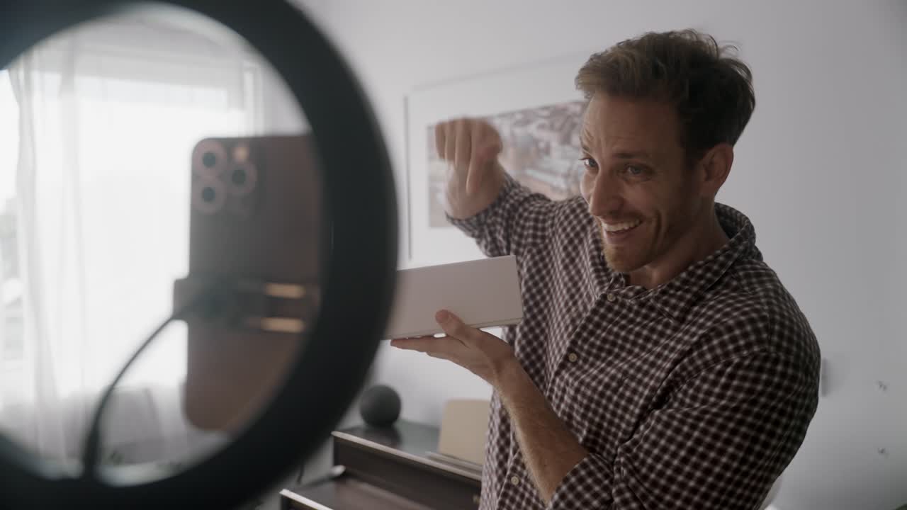 Homem gravando vlog ou reel para redes sociais, analisando um produto em vídeo de unboxing transmitido ao vivo. Influenciador estilo UGC falando ao celular em estúdio caseiro. Criador de conteúdo animado e feliz abrindo caixa branca.