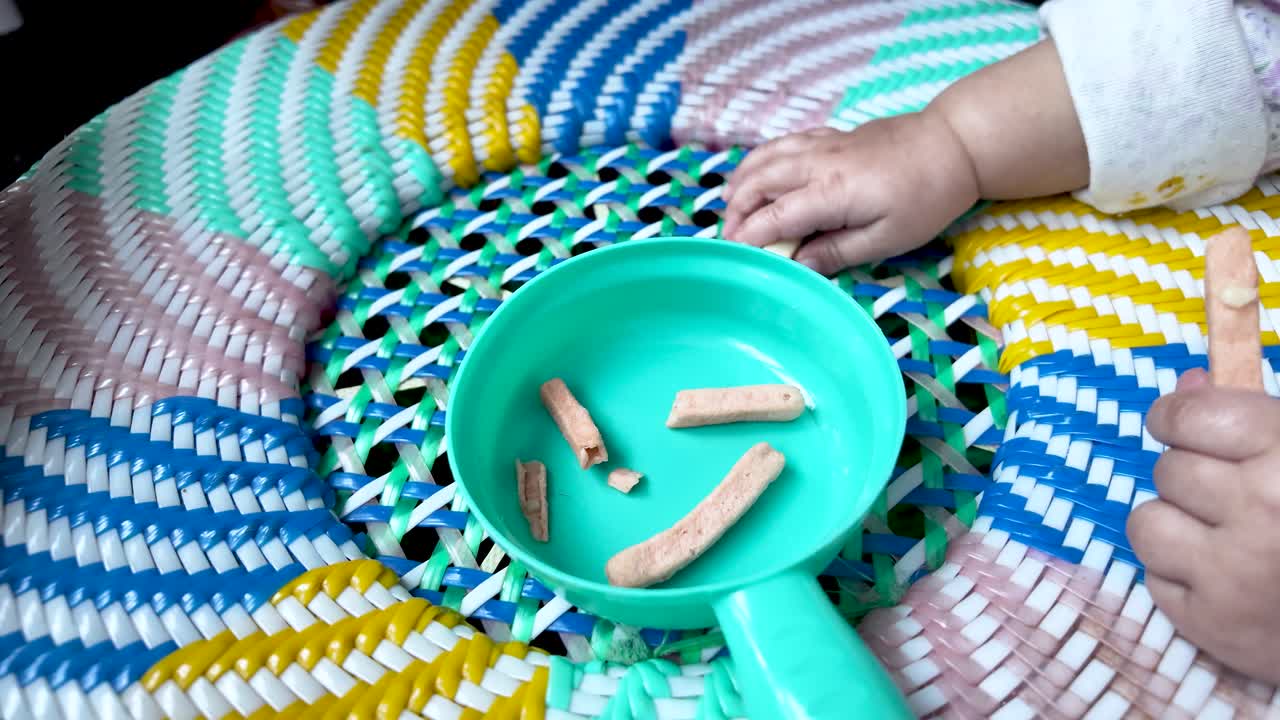Baby greift nach Gemüsesticks im blauen Spieltopf auf einem bunten, gewebten Rohrstuhl. Gesunder Snack für Kleinkinder, Ernährung in der frühen Kindheit, Spielzeit