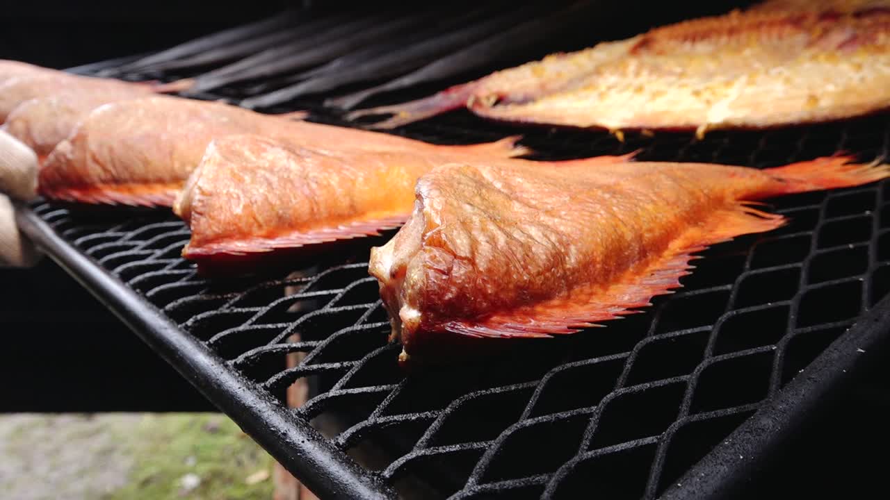 Various types of fish, including eel and redfish, are being smoked on a grill rack. The golden-brown color and rich texture highlight the traditional smoking process.