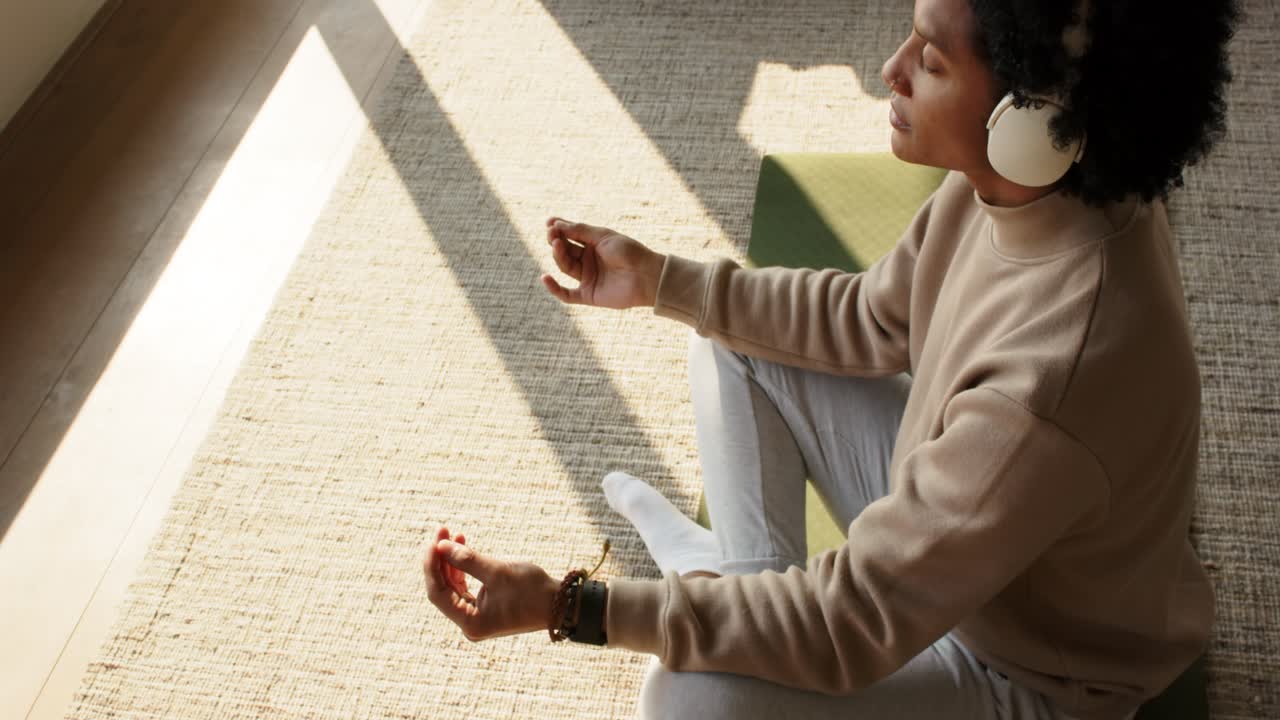 Man sitting on rug, unfolding yoga mat, wearing headphones, meditating at home for calm, copy space