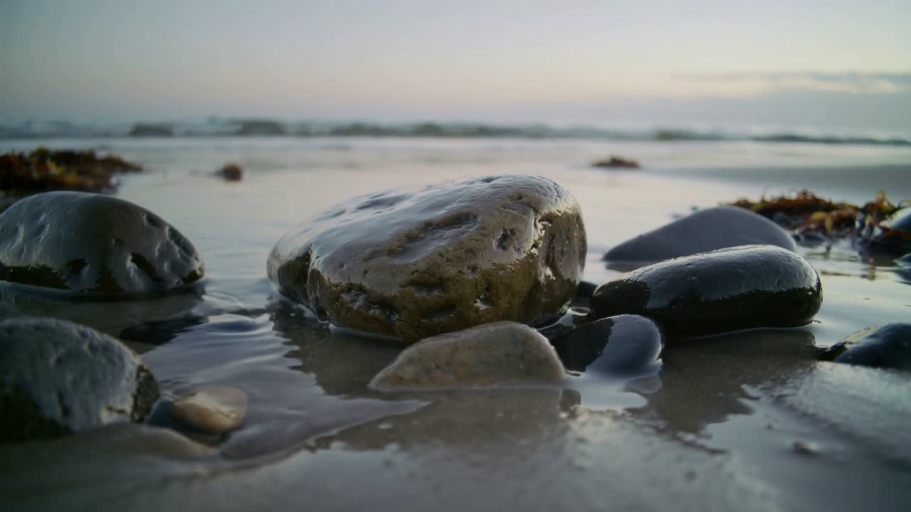 Acqua di mare bassa che avanza spingendo dolcemente sassi lisci e bagnati sulla battigia, formando riflessi lucidi.