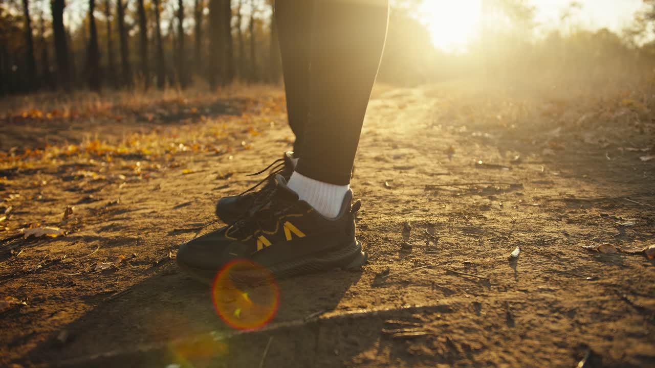 de cerca, un hombre con un uniforme deportivo negro y zapatillas negras estira sus pies antes de comenzar a correr a lo largo de un camino de tierra en el bosque de otoño al amanecer en un soleado día de otoño