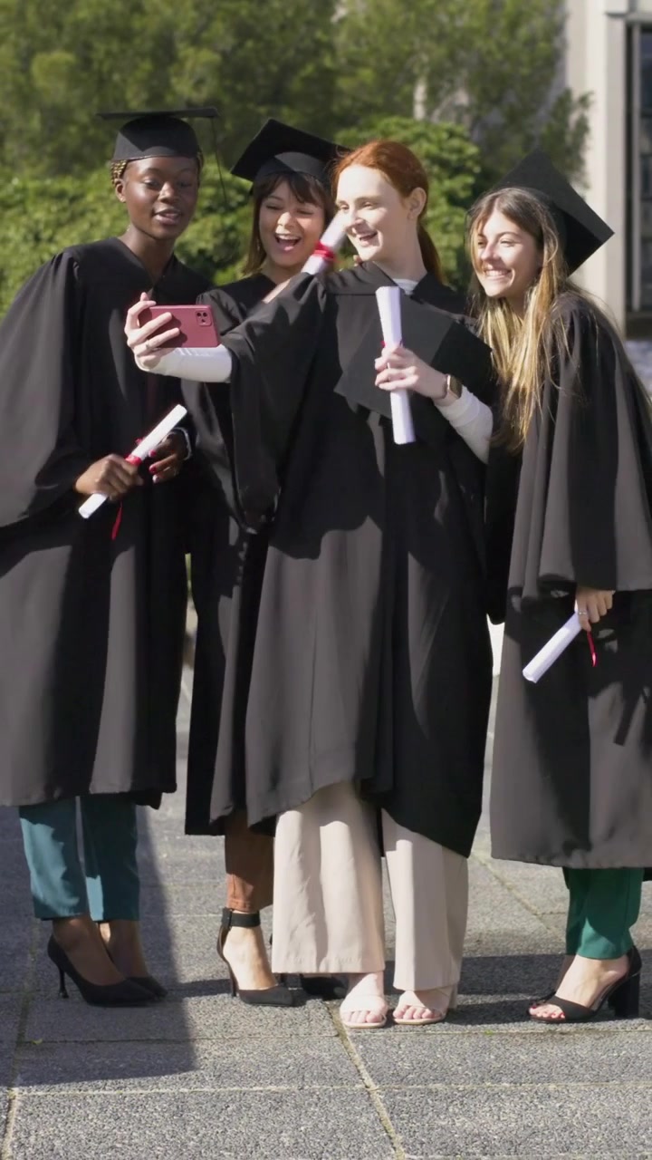 Vertical video: Extending pink phone, grad taking selfie peers joining on campus in gowns, scrolls