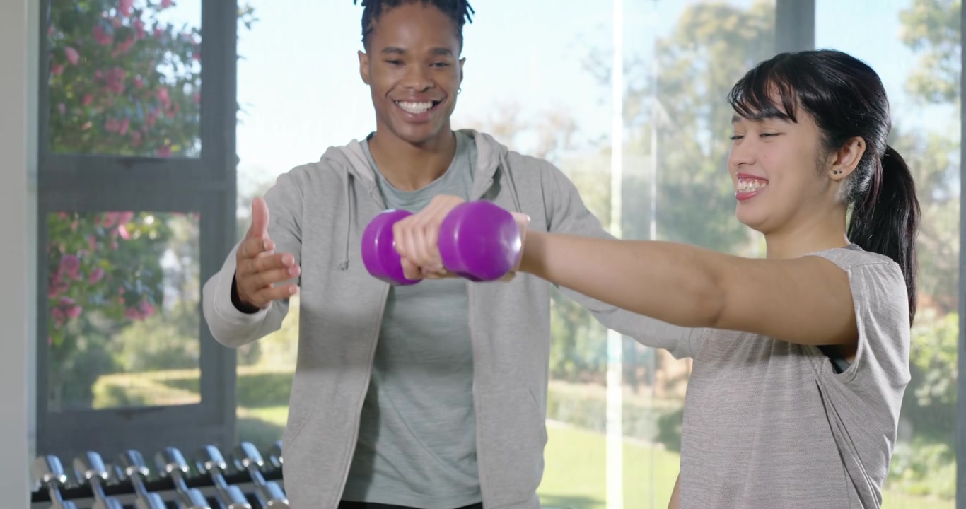 Male trainer guiding Asian trainee doing front raises with purple dumbbell in gym, correcting form