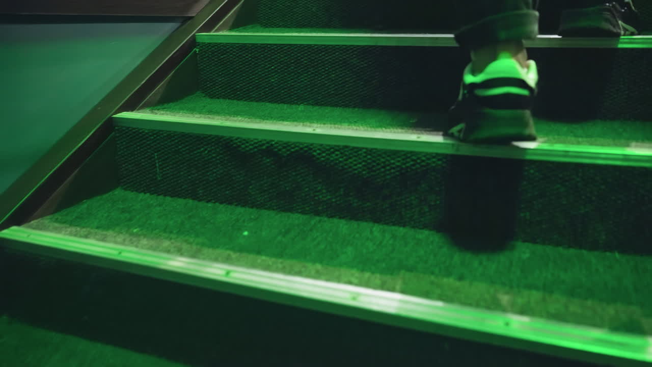 검은색과 색의 스니커를 입은 발이 녹색 조명 아래 카으로 된 계단을 올라 빌리어드 방을 향해 습니다. 금속 레일링은 깊이와 현대적인 느낌을 더합니다. 근접 촬영은 움직임과 질감을 포착합니다.