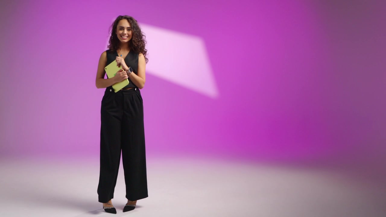 Woman with curly hair holds a yellow tablet on a purple studio background, looking at the camera, smiling and posing confidently while wearing an elegant black outfit in a professional fashion scene