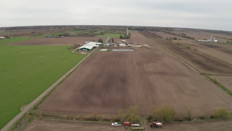 el paisaje agrícola plano con vista aérea desde el dron que se aleja del granero y el silo revela campos recién arados, camiones y tractores trabajando para prepararse para la temporada de siembra de primavera