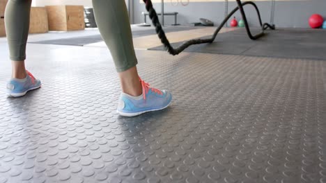 sección baja de una mujer biracial inalterada haciendo ejercicio con cuerdas de lucha en el gimnasio, en cámara lenta.