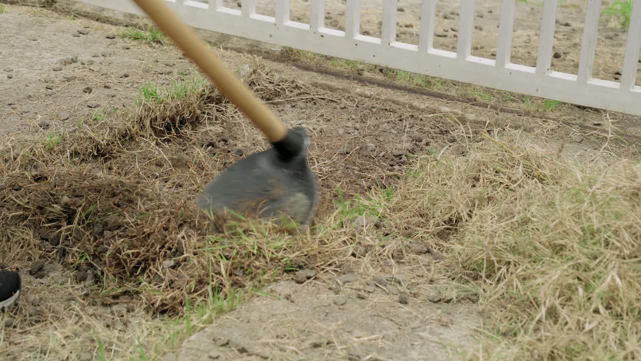 High angle shot of a person using a gardening hoe to clear weeds, dry grass, and till the dirt ground next to a white fence in a backyard