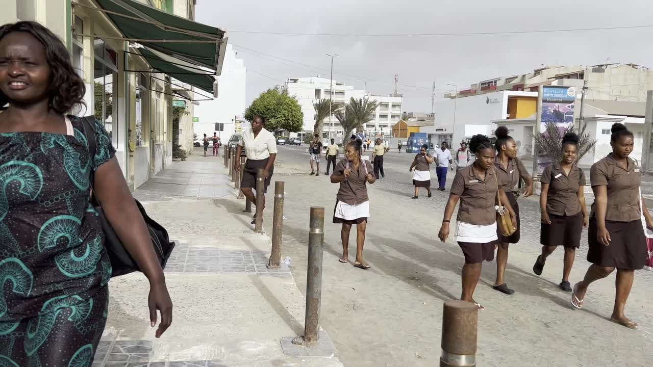 First-person view of Creole workers walking along sidewalk in Sal Rei, Cape Verde, heading home after shift. The bustling street captures local life, urban charm, and vibrant community dynamics