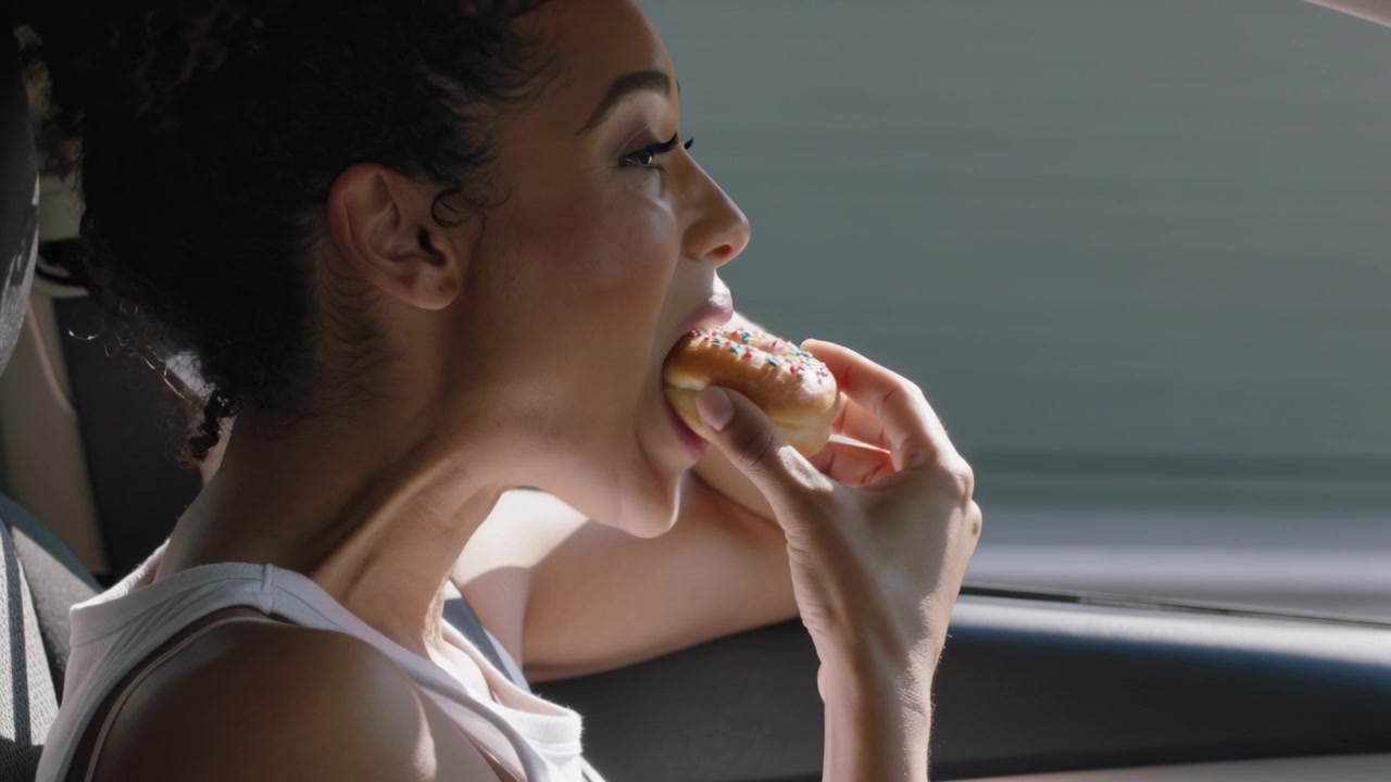 Holding sprinkled doughnut, woman in white tank top inspecting and biting for snack in moving car