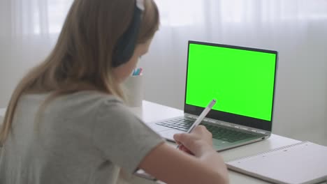 una adolescente está aprendiendo en línea sentada en la mesa frente a una computadora portátil con la tecla chroma en exhibición, escribiendo en un cuaderno en casa