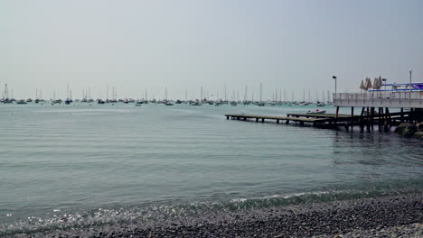 static shot of wooden pier along the shoreline with sail boats docked in distance in la punta, callao, peru on a sunny day