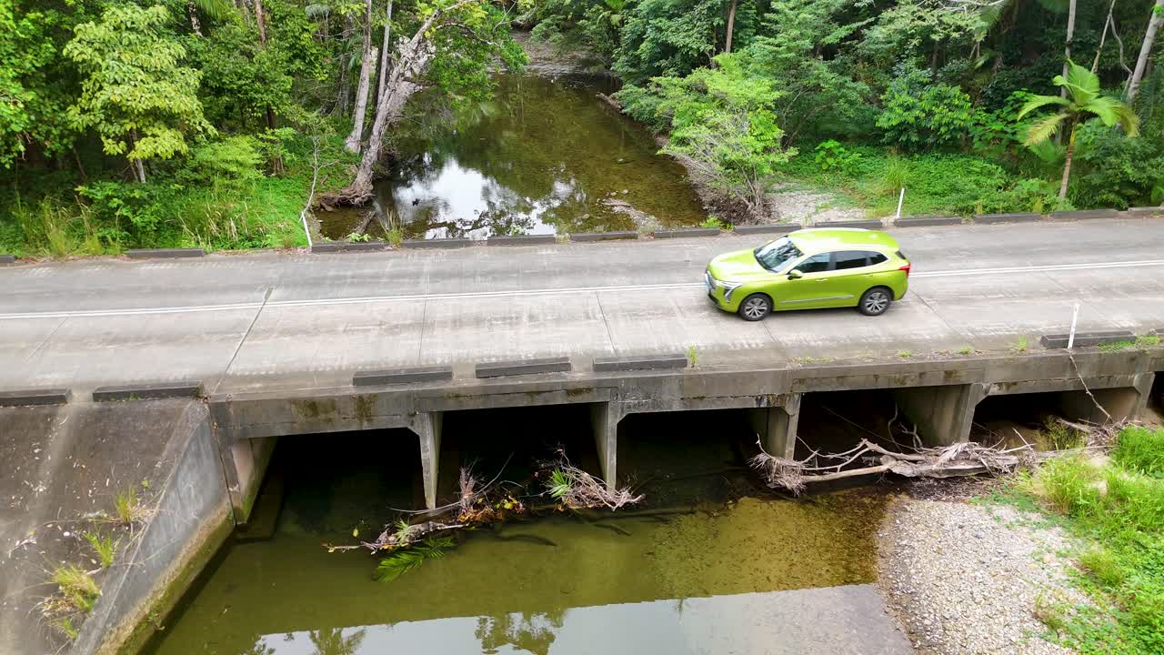 A green car drives across a bridge over a creek in a lush rainforest setting, captured by a drone