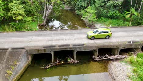 a green car drives across a bridge over a creek in a lush rainforest setting, captured by a drone