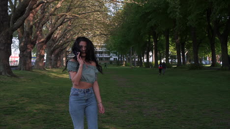 shot from behind of an attractive and playful latina woman with black wavy hair walking under the trees in a park in london, turning and looking at the camera, happy with a beautiful smile