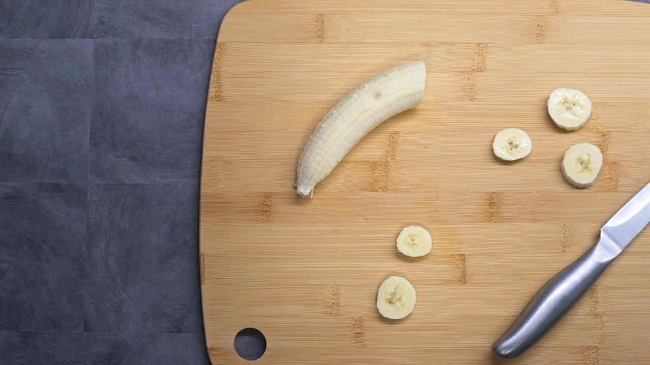 A partially peeled banana lies on a wooden cutting board with several freshly sliced pieces beside it. A knife is placed nearby, indicating food preparation.