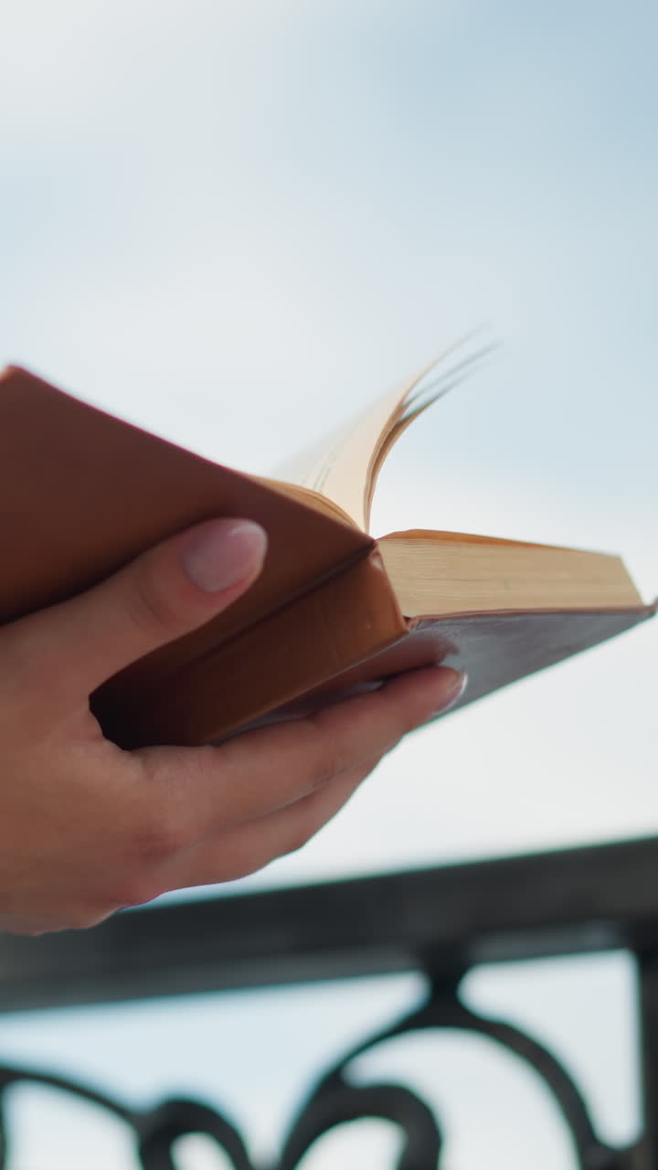 Closeup of a lady holding an open book in her right hand, with her left hand supporting the book from beneath, she stands close to an iron fence, with the pages slightly lifting as if from a breeze