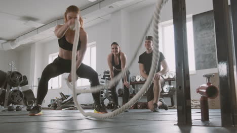 culturista de crossfit trabajando con cuerdas de combate mientras sus compañeros de gimnasio la apoyan 1