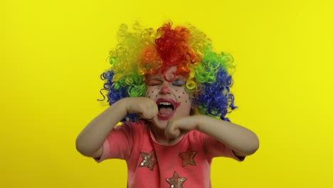 Little-child-girl-clown-in-colorful-wig-making-silly-faces,-crying.-Halloween.-Yellow-background