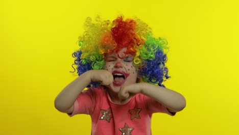 little child girl clown in colorful wig making silly faces, crying. halloween. yellow background