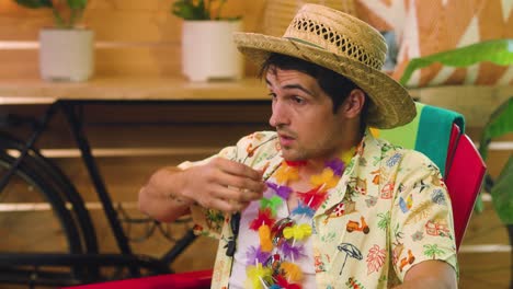 Un-Joven-Vestido-Con-Un-Traje-Tropical-Hawaiano-De-Vacaciones,-Relajación,-Playa,-Traje-Lei,-Sombrero-Para-El-Sol,-Se-Sienta-Durmiendo-En-El-Trabajo,-Se-Despierta-En-Una-Silla-De-Madera-Roja-Dentro-De-Una-Tienda-De-Muebles-Durante-El-Día