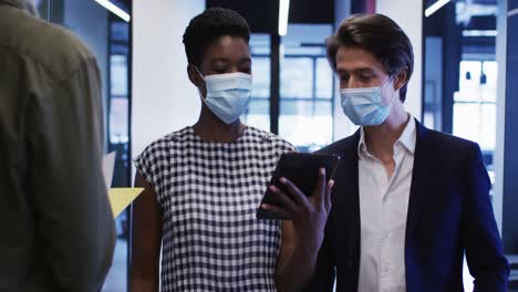 diverse business people wearing face masks talking using digital tablet walking through office