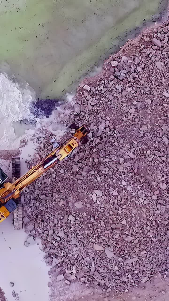 Excavator moves stones and rubble from river edge onto pile in construction zone, slow motion