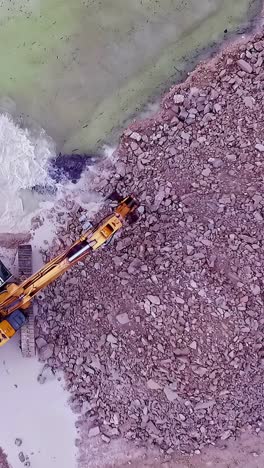 excavator moves stones and rubble from river edge onto pile in construction zone, slow motion
