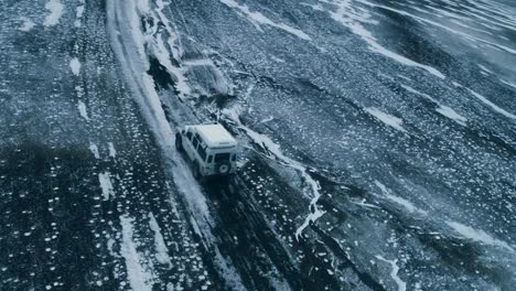 aerial drone footage of a white 4x4 truck vehicle car driving on ice and snow covered rural and desolate road in the mountains at greenland or iceland during a cloudy day with snow covering the ground