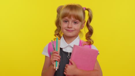 cheerful funny schoolgirl kid with books dressed in uniform wears backpack smiling looking at camera