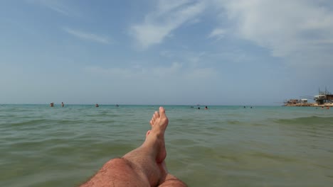 first-person view of male tourist legs relaxing and floating on inflatable mattress with trabucchi or trabocchi and people bathing in background