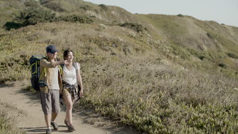 happy father and daughter walking along dirt road in mountains, carrying backpacks