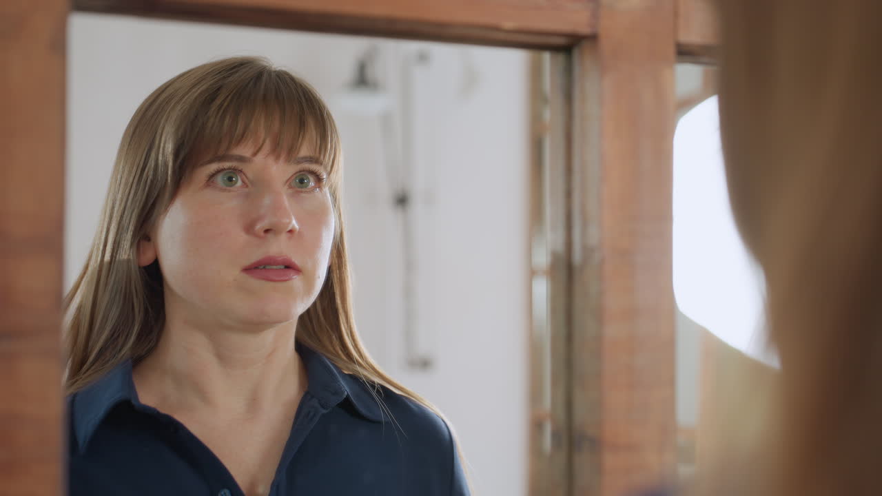 Mirror reflection of young lady wearing blue as she arranges her face with elegance, gestures confidently while smiling and winking softly at herself, engaging in calm self care ritual indoors