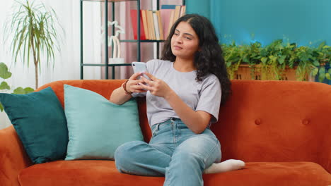 Indisch-arabische-Junge-Frau,-Die-Auf-Dem-Sofa-Sitzt,-Nutzt-Ihr-Smartphone-Und-Teilt-Nachrichten-In-Einer-Social-Media-Anwendung