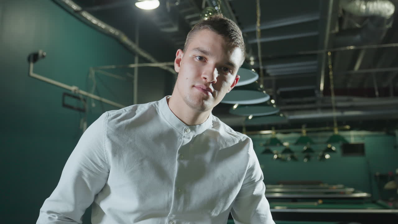 색 셔츠를 입고 약간 미소 짓는 남자가 희미하게 밝은 빌리어드 홀에서 큐 스틱을 들고 있는 클로즈업. 오버헤드 조명은 초록색 빌라 테이블에 반사를 던집니다. 자신감있는 표정은 집중과 전략을 암시합니다.