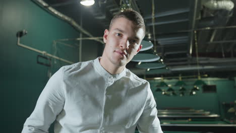 close-up of man in white shirt with slight smile holding cue stick in dimly lit billiards hall. overhead lights cast reflections on green pool table. confident expression suggests focus and strategy