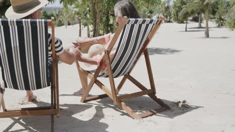 Happy-senior-caucasian-couple-sitting-in-deckchairs-on-sunny-beach-holding-hands,-in-slow-motion