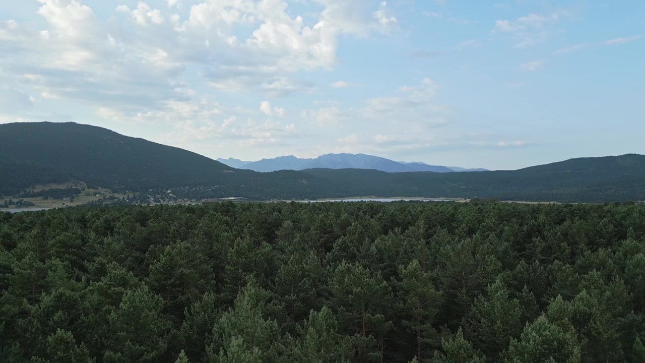 Camera pans over a lush green forest revealing the vast expanse of lac de matemale and a breathtaking mountain range in the distance