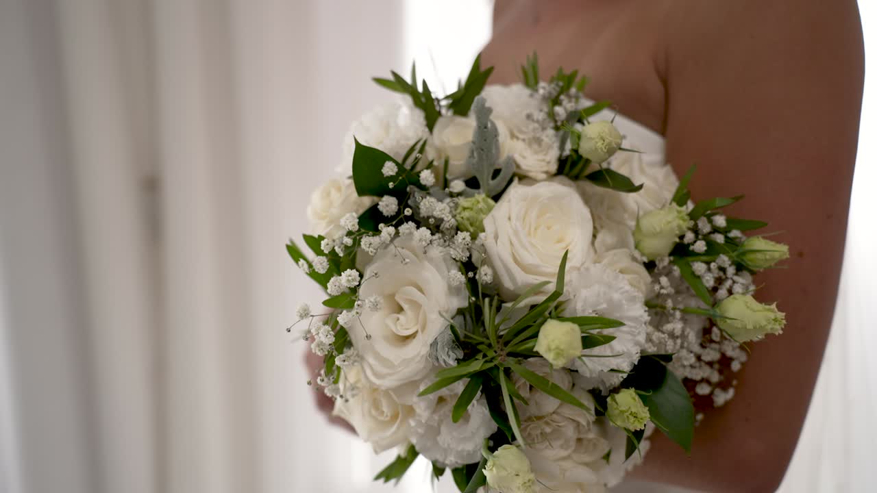 Una sposa, in una stanza luminosa, tiene in mano un bouquet di fiori bianchi. L'attenzione è focalizzata sulle sue mani e sulla bellissima composizione floreale mentre si prepara per la cerimonia.