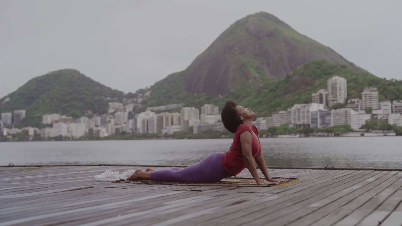 A woman practices yoga on a wooden pier overlooking a lake with a city and mountains in the background