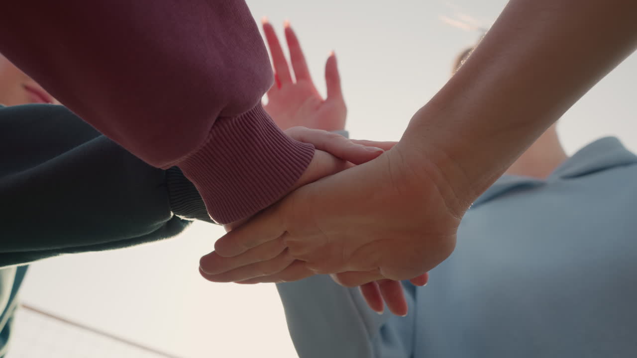 후디를 입은 사람들이 팀원으로서 손을 함께 놓고 빛이 손을 둘러싸고 빛나는 효과를 일으켜 야외 환경에서 단결과 팀워크를 상징합니다.