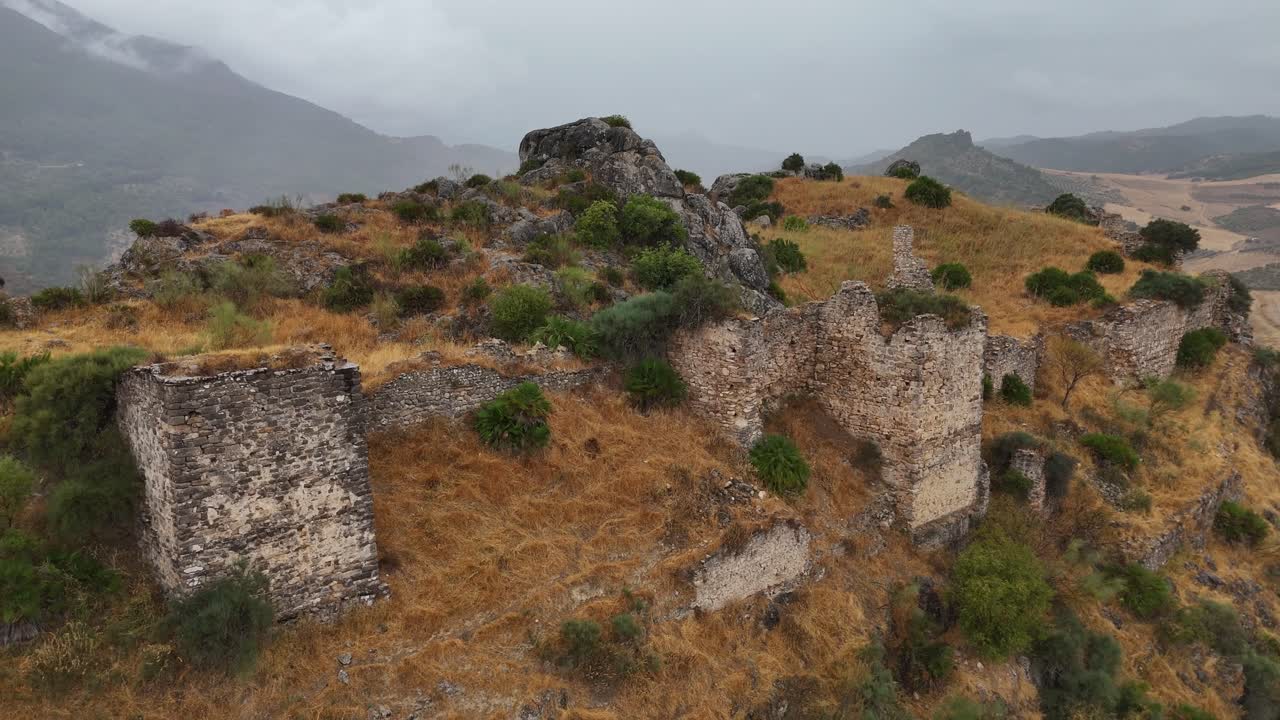 aerial view tracking back from Turón Castle ruins, Ardales, Málaga, Spain, revealing stone walls and the surrounding farmland under an overcast sky