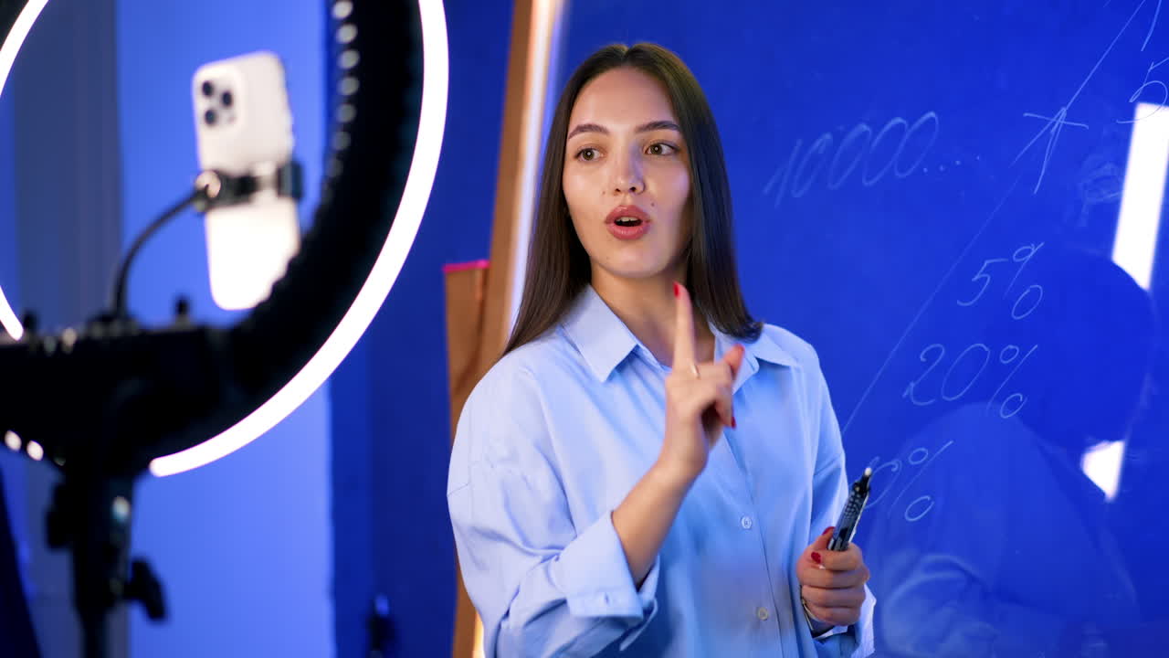 Long-haired woman stands at the glass wall and writes with white marker. Female blogger streaming using a phone camera on tripod with light ring.