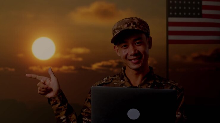 Primo piano di un soldato asiatico che usa un laptop e indica di lato, in piedi con la bandiera degli Stati Uniti, al tramonto