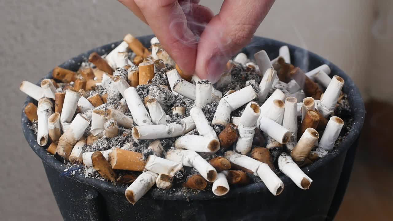 Slow-motion footage of an ashtray full of cigarettes and a hand erasing a cigarette, with some smoke.