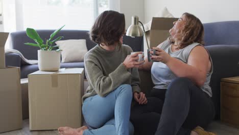 caucasian happy lesbian couple drinking tea sitting on floor in new home